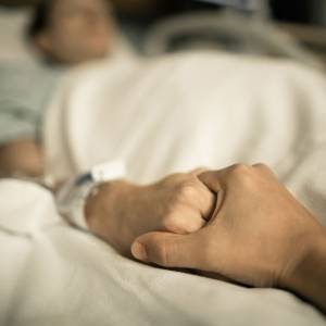 A person holds the hand of a dying patient in a hospital bed.