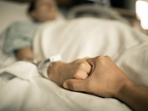 A person holds the hand of a dying patient in a hospital bed.