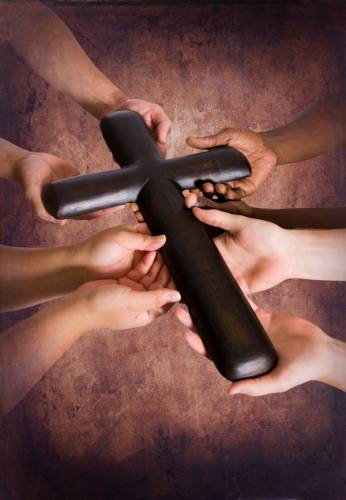 Several hands of people of different ethnicities hold a small wooden cross