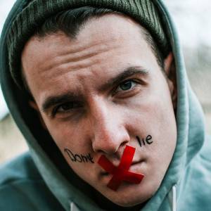 Man with red X taped to his lips with the words “Don’t lie” written on his face.