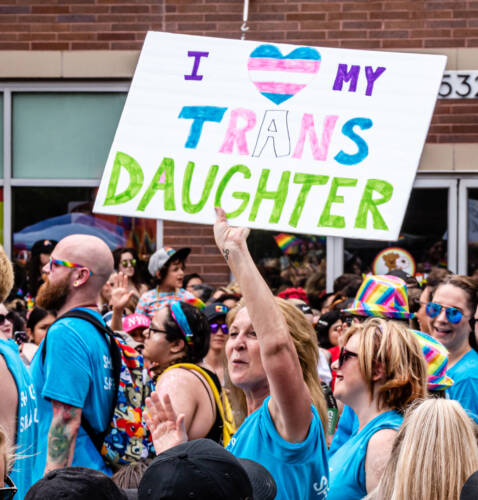 A woman in a LGBTQIA+parade holds a sign saying, "I heart my trans daughter."