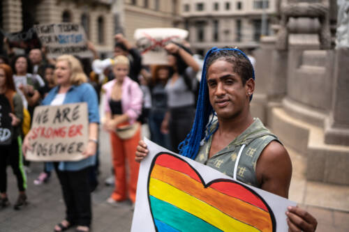 Transgender woman of color with an electric blue weave holds a rainbow heart at a demonstration. A middle aged white woman with blonde hair wearing a denim shirt, brown pants and sensible shoes holds a cardboard sign that reads, "We are Stronger Together." 30 others are in the field of visions also with signs. The setting is of marble buildings and an ornate marble staircase or fountain in the back right corner.