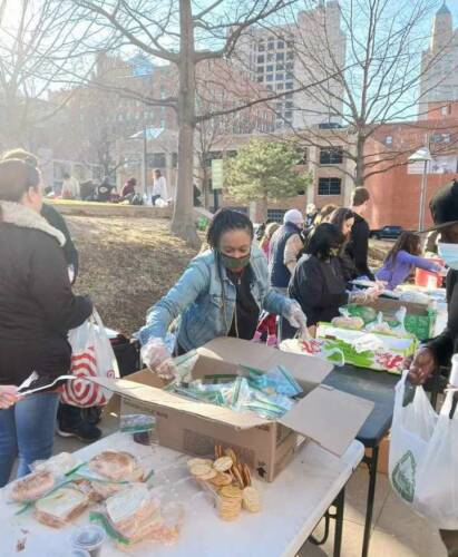 Photo of tables with food set up outdoors with city park and skyline in background. Church members serving others packed lunches.